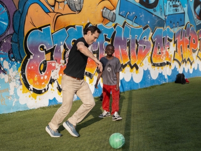 An adult man plays soccer with a young boy in front of a colorful graffiti mural