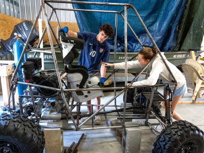 Two students work inside a workshop on a custom Baja-style vehicle frame, with one pointing inside the chassis while the other positions a metallic racing seat.