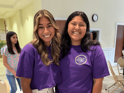 Two smiling young women stand together wearing matching purple t-shirts and white pants in a brightly lit indoor event space.