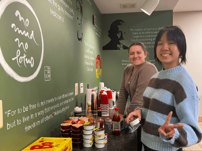 Two smiling women stand at a food counter with Bojangles boxes and trays of food, in front of a green wall featuring quotes and stencil art.