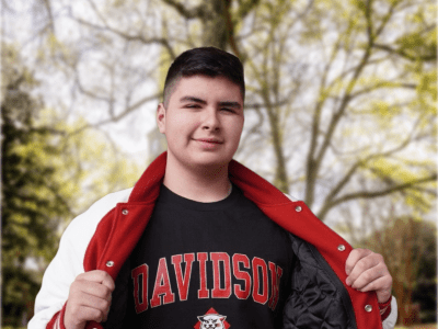 A young man smiles while holding open a red and white letterman jacket to reveal a black t-shirt featuring the word "DAVIDSON" in red block letters and a wildcat mascot logo, set against a blurred outdoor campus background.