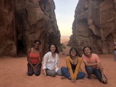 Four smiling students sit together on pink desert sand framed by towering rock canyon walls.
