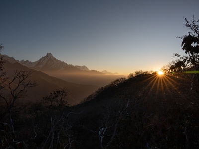The sun rises over a dark, silhouetted hillside, casting bright starburst rays across a misty valley toward a jagged, snow-dusted mountain peak in the distance.