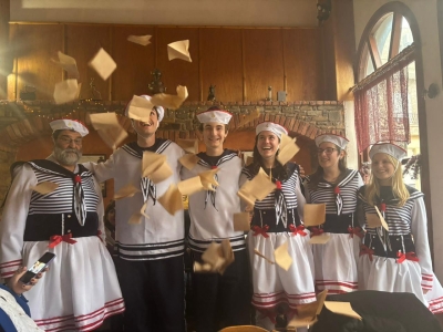 Six people dressed in matching sailor costumes smile and laugh indoors as square pieces of tan paper rain down around them.