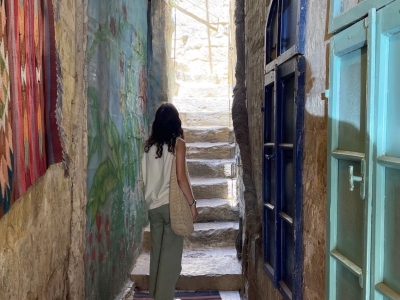 A person stands looking up a narrow stone staircase at the end of an alleyway decorated with a colorful rug, a painted wall mural, and vibrant window frames.
