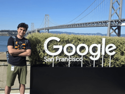 A man stands with crossed arms next to a large "Google San Francisco" sign on an outdoor patio, with the San Francisco-Oakland Bay Bridge stretching across the background under a clear blue sky.