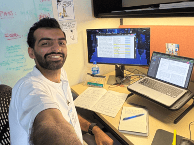 A smiling man takes a selfie at his office desk, which features a laptop on a stand, an external monitor displaying a spreadsheet, notebooks, and a nearby whiteboard with text.