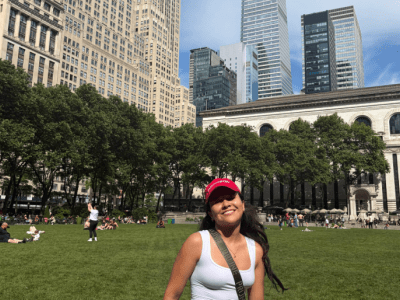 A smiling young woman wearing a white tank top, denim shorts, and a red baseball cap stands on a large green lawn in front of tall city skyscrapers and a historic building under a blue sky.