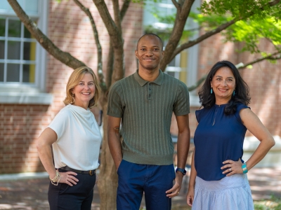 Three diverse colleagues smile at the camera while standing together outdoors in front of a tree and brick building.