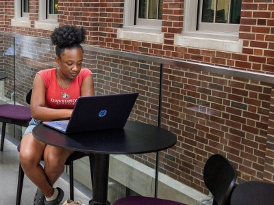 A female student in a red Davidson College shirt works on a laptop at a small round table next to a brick wall.