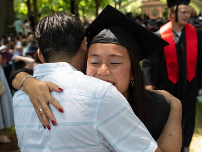 a young woman in graduation regalia hugs another person on a sunny graduation day