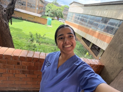 A smiling woman in blue medical scrubs takes a selfie outdoors in front of a red brick walkway and multi-story institutional buildings surrounded by green lawn and trees.