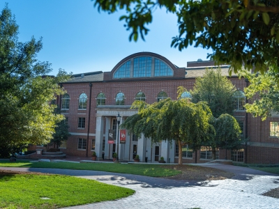 Alt Text:  Sunlight illuminates the brick exterior and columned entrance of Alvarez College Union, framed by green trees and paved campus walkways on a clear day.