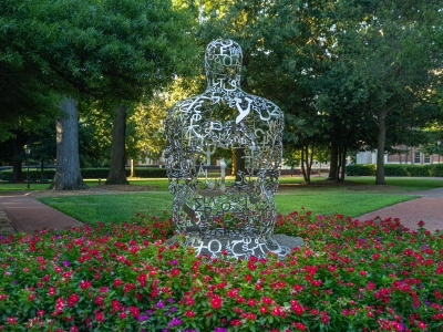 A hollow metal sculpture of a human figure crafted from international letters and symbols sits surrounded by a bed of vibrant pink flowers on a green campus lawn