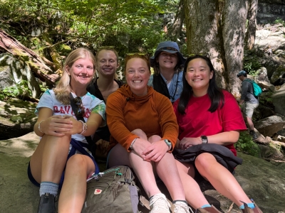 Five smiling friends sit together on large rocks outdoors during a trail hike in a lush, wooded forest.   