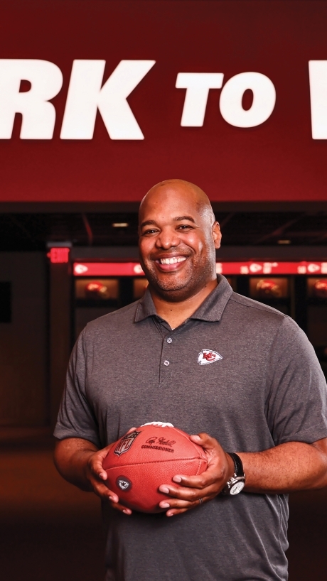 a young Black man holding a football in front of a wall that reads "work to win"