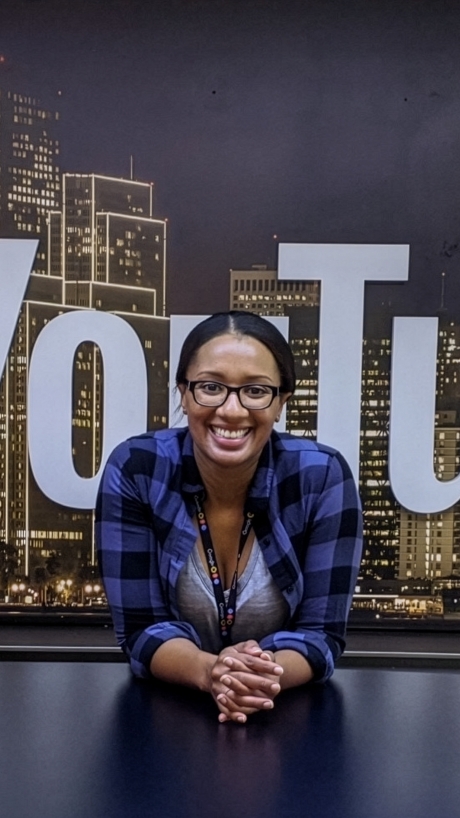 A smiling woman with elbows on a newsroom desk and the YouTube logo behind her