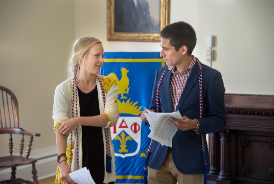 Two students stand in front of a colorful flag for Pi Delta Phi talking with honors cords around their necks