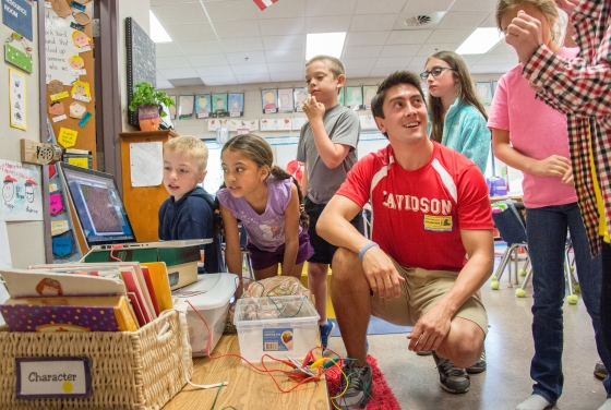 Davidson student wearing a Davidson soccer jersey stands amidst a group of young kids in their classroom and shows them math exercises on a laptop
