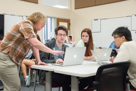 Professor Heyer stands near table and talks to three students who are using laptops in her class