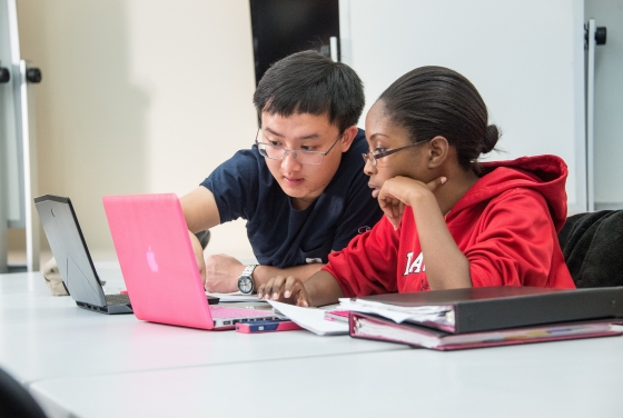 Two students talk with laptops open, one pointing at his colleague's laptop