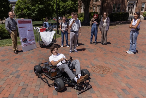 A group of students and faculty stand around and smile as a student drives a go-kart
