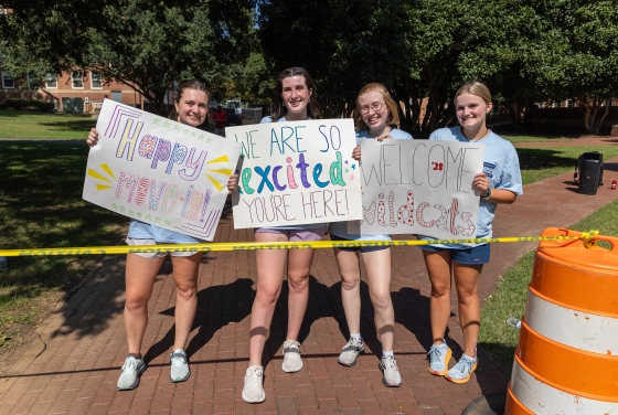 a group of four young women hold signs "welcome" to campus