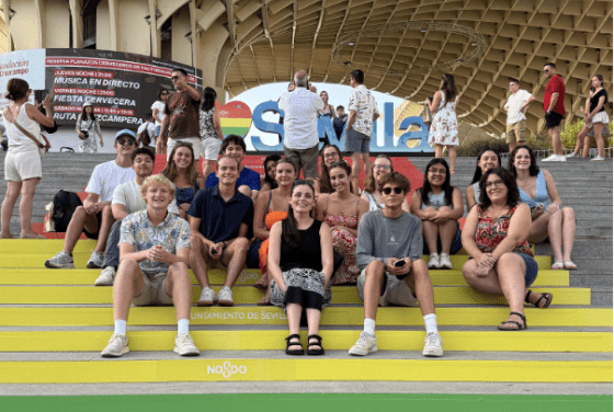 A group of approximately fifteen students sits on yellow and green steps branded with "Ayuntamiento de Sevilla" and "NO8DO" logos, positioned in front of the Setas de Sevilla wooden structure and a colorful "Sevilla" sign.