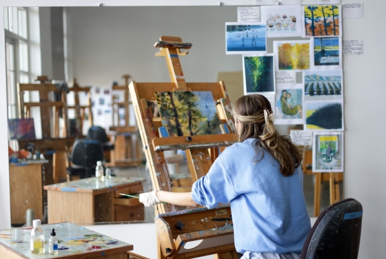 An art student in a light blue sweatshirt sits at a wooden easel painting a landscape, with their workspace and various reference images reflected in a large studio mirror.