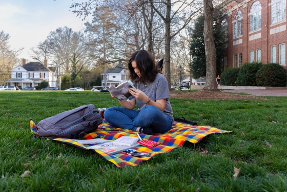 A student sits cross-legged, reading a book, on a rainbow picnic blanket on a green lawn