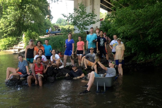 A large group of diverse young adults wearing work gloves poses for a photo while standing and sitting in a shallow river and on rocky banks beneath a concrete bridge overpass.