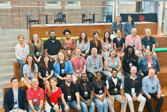 A diverse group of approximately twenty-five people sit on wide, tiered wooden bleachers indoors for a large group portrait.