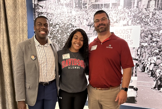 Three smiling people pose for a photo in front of a historic black-and-white mural of a crowded street, with the woman in the center wearing a gray sweatshirt that says "Davidson Alumni."