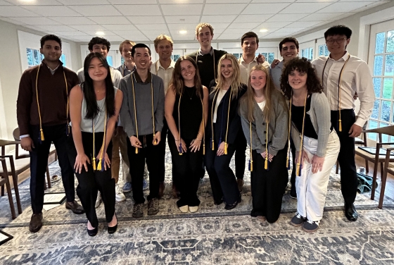 A large group of students, many wearing blue and gold honor cords over their shoulders, pose together for a group photo in a brightly lit room.