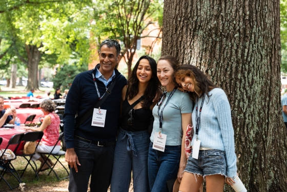 A smiling family of four poses together under a large tree during a Davidson College orientation event, with a background of outdoor tables and green foliage.