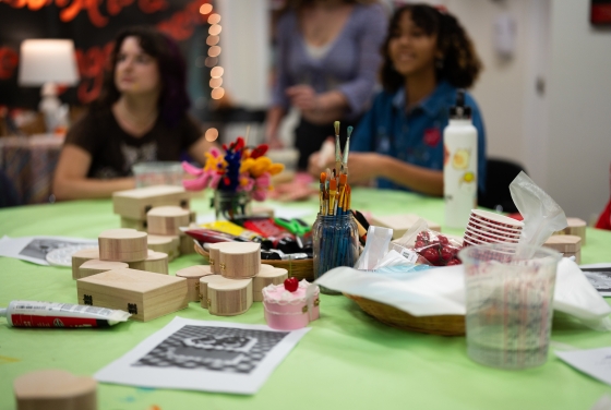 A green table is covered in art supplies, including unfinished wooden heart-shaped boxes, jars of paintbrushes, and acrylic paint tubes, while students gather in the blurred background of the studio.