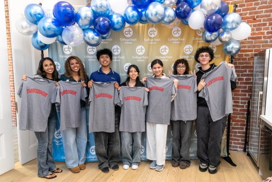 Seven smiling students stand in a row in front of a College Track Oakland backdrop and a blue and white balloon arch, each holding up a grey T-shirt with the word "DAVIDSON" printed in red arched lettering.