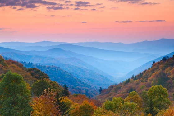 A scenic overlook of rolling mountain ridges receding into a hazy blue distance under a vibrant sunset sky of pink and orange clouds, with a foreground of lush autumnal trees in shades of green, gold, and rust.