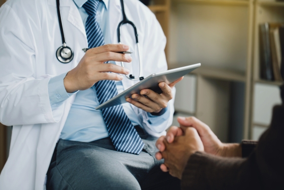 A male doctor wearing a white lab coat, stethoscope, and blue striped tie holds a digital tablet and pen while speaking with a patient whose hands are clasped in the foreground.