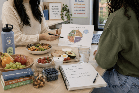 A dietitian points to a "Building a Balanced Plate" diagram while consulting with a client at a desk covered in healthy food items, nutrition books, and a notebook listing health questions.