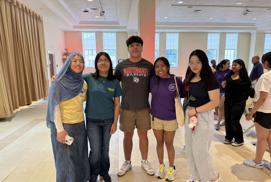 Five smiling college students pose together in a brightly lit indoor venue, with the student in the center wearing a charcoal grey Davidson Wildcats t-shirt.