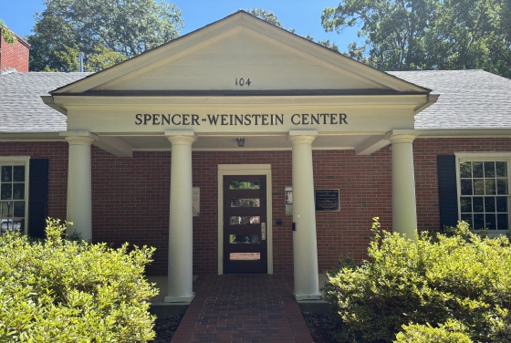 The exterior front entrance of the red brick Spencer-Weinstein Center building, featuring four white columns, a triangular pediment, and a brick walkway lined with green bushes.