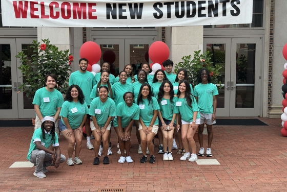 A diverse group of student leaders wearing matching teal t-shirts smiles and poses together on a brick courtyard underneath a large banner reading "WELCOME NEW STUDENTS."