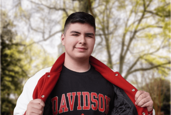 A young man smiles while holding open a red and white letterman jacket to reveal a black t-shirt featuring the word "DAVIDSON" in red block letters and a wildcat mascot logo, set against a blurred outdoor campus background.