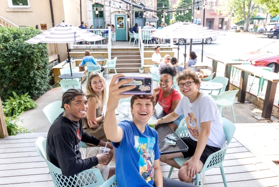 Five smiling students sit together around a patio table outdoors while one takes a group selfie with a smartphone.