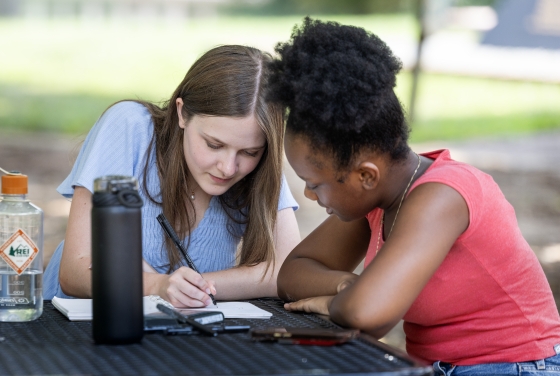 Two young women sitting at an outdoor table studying or writing together in a notebook.
