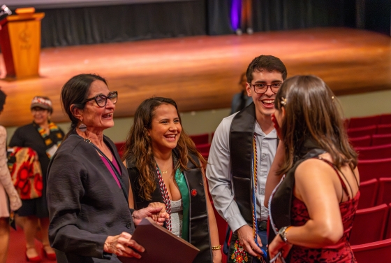 A group of smiling graduates wearing graduation stoles talk and laugh with a professor in an auditorium.