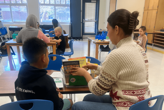 A instructor in a patterned sweater sits across a table from a young student in a classroom, selecting books from a green bin for their reading activity.