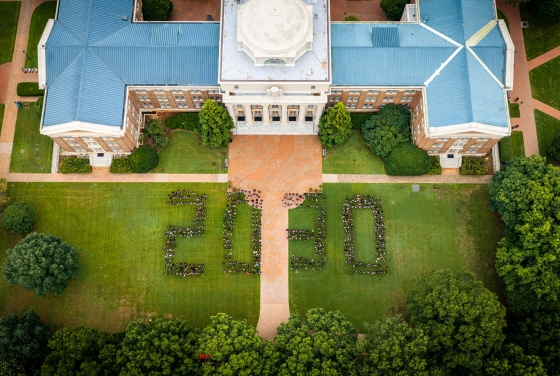 Aerial view of a large group of students standing on a green lawn in front of a domed brick campus building, forming the numbers "2030."