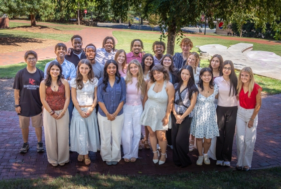 A large group of young adults smile together while standing outdoors on a brick pathway in front of a grassy lawn with trees.